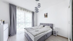 Une chambre moderne avec un lit recouvert d’un tissu gris, une table de chevet avec une lampe, une petite table de nuit blanche et une fenêtre avec des rideaux gris.