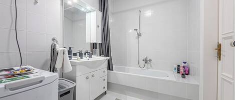 Un baño con lavamanos blanco, espejo y ducha. Hay un perchero, un cubo de basura y artículos de aseo en la encimera.