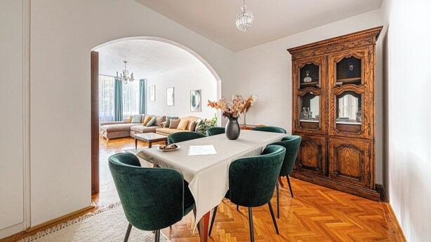 Een eetgedeelte met een tafel voor vier personen, groene fluweelstoelen, een houten kast met glazen deuren en een kroonluchter.