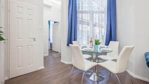A dining area with a round glass table, white chairs, and a window with blue curtains. - Quiet Downtown Haven— 2Br | A/C | Free Parking (Budapest)