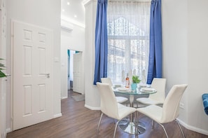 A dining area with a round glass table, white chairs, and blue curtains. - Quiet Downtown Haven— 2Br | A/C | Free Parking (Budapest)