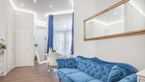 A modern living room with a blue velvet sofa, white walls, and a large mirror. - Quiet Downtown Haven— 2Br | A/C | Free Parking (Budapest)