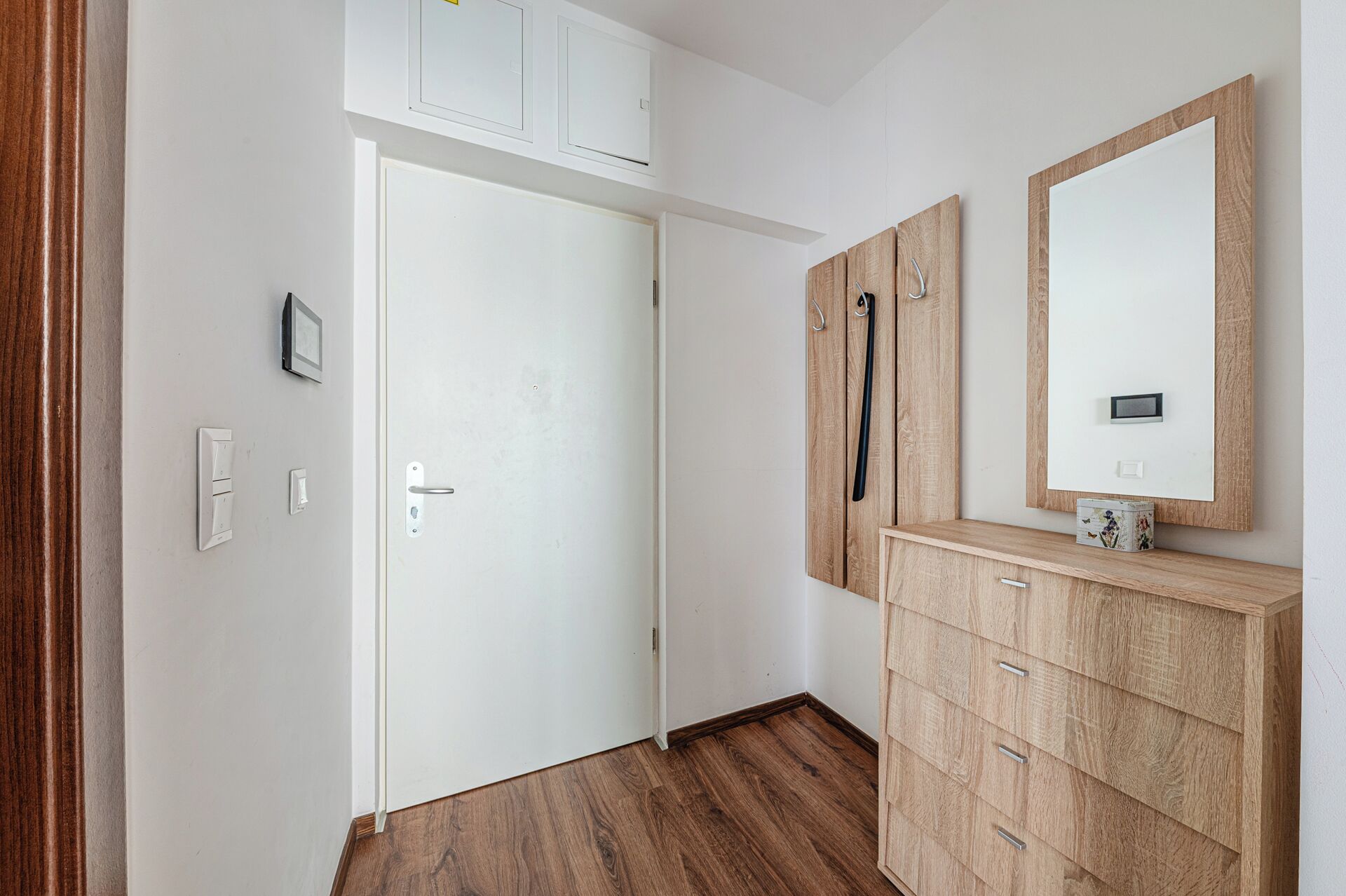 Een kamer met een houten vloer, een witte deur, een houten garderobe met spiegel en een klein fotolijstje aan de muur.