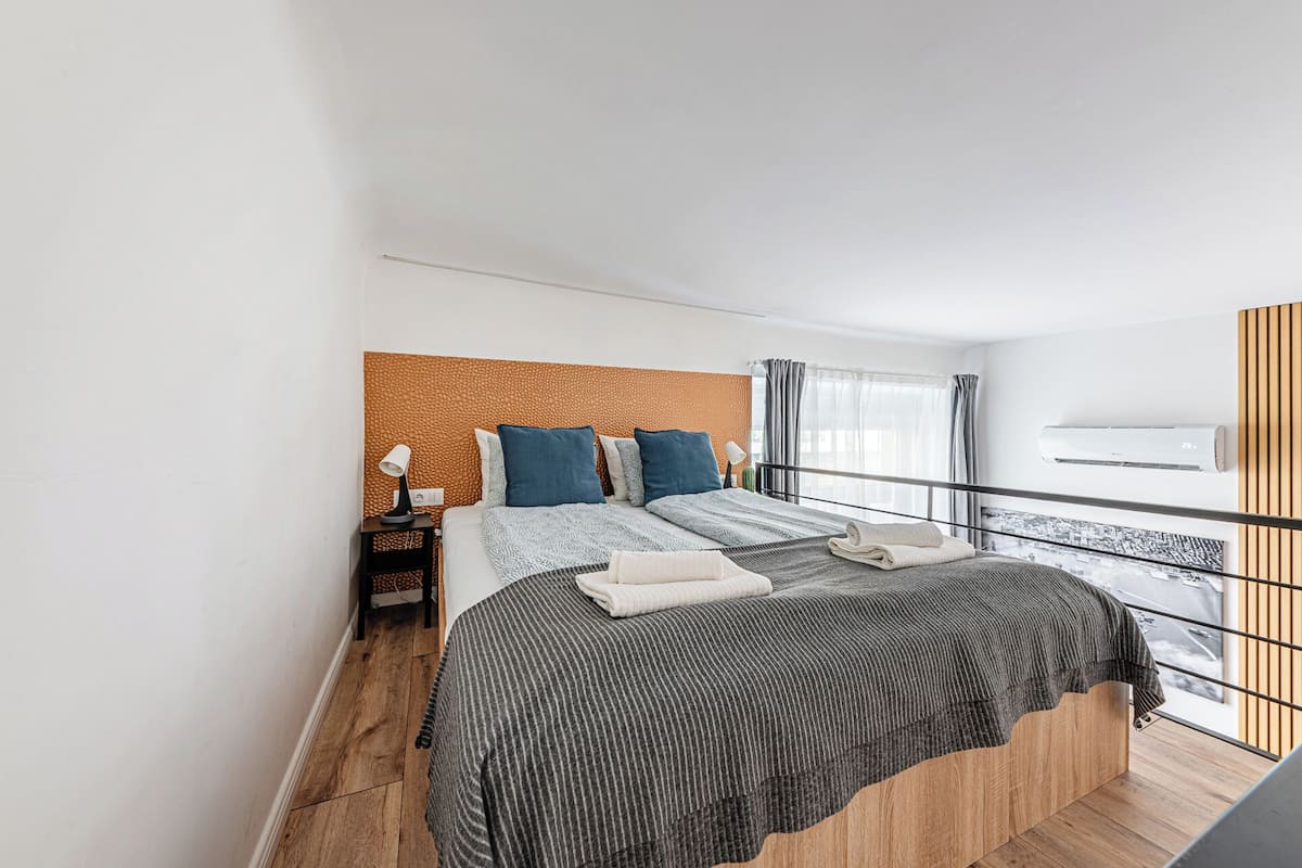 A modern bedroom with a wooden headboard, a large bed with striped bedding, a nightstand with a lamp, and a balcony with a railing.