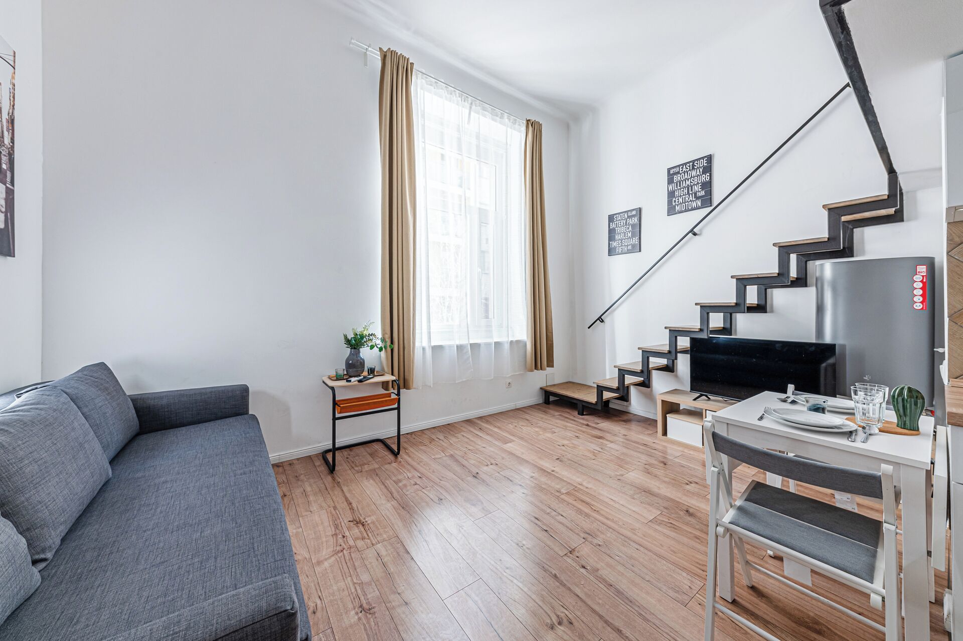 A modern living room with a grey sofa, a small side table with a plant, a staircase with a TV, and a wall with a sign.