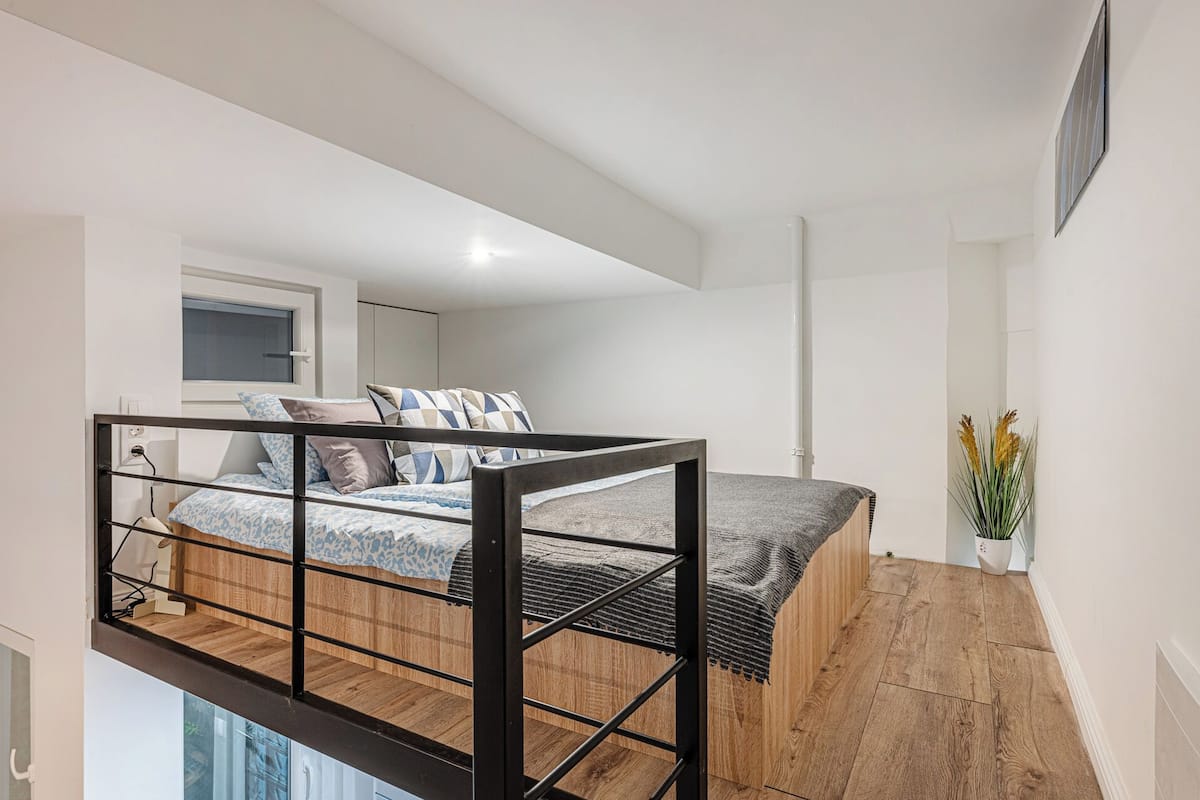 A modern loft bedroom with a bunk bed, wooden flooring, a small window, and a potted plant.