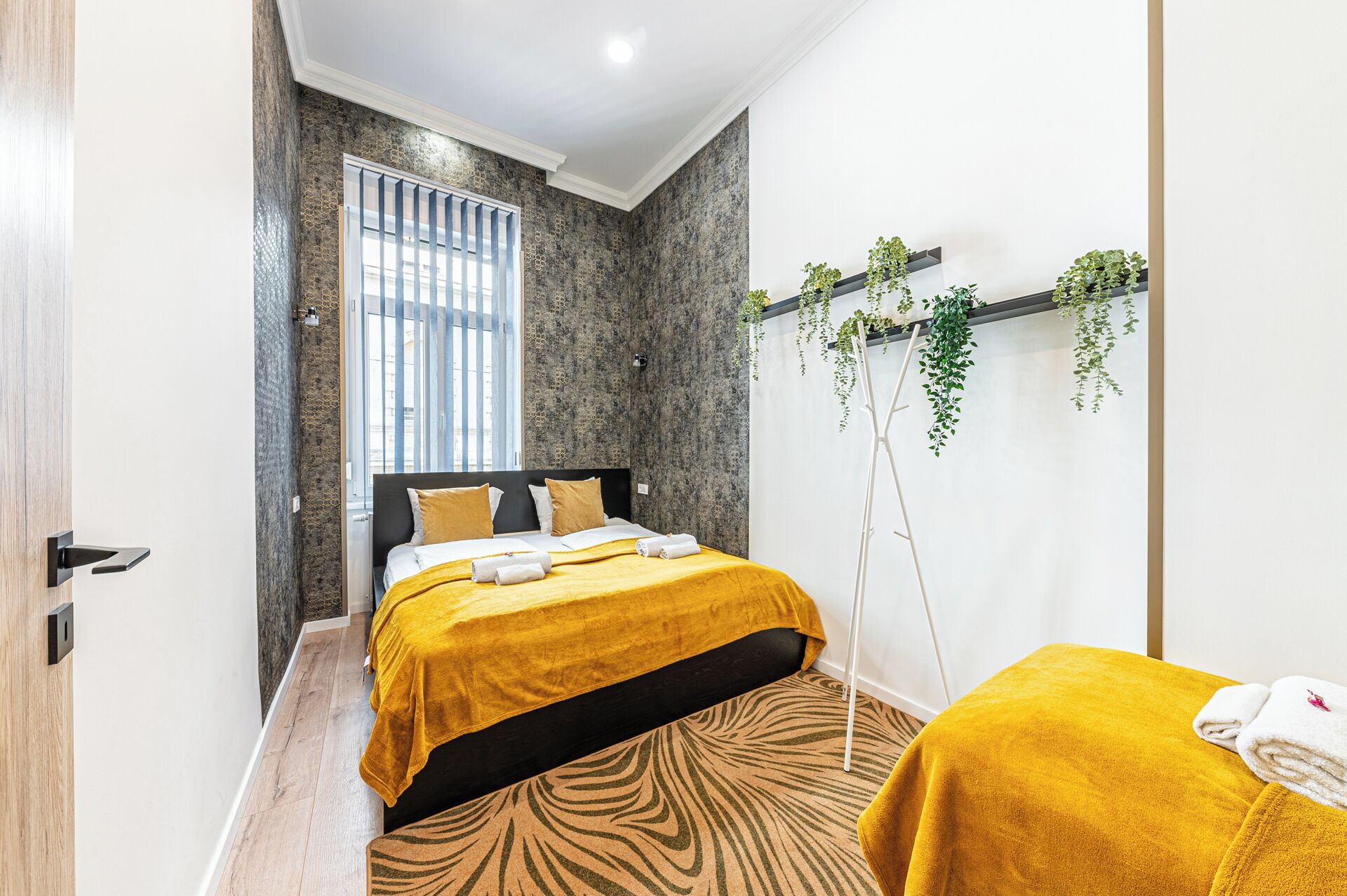 Habitación de hotel con cama, ventana con persianas, alfombra de estampado de cebra y planta en un estante.