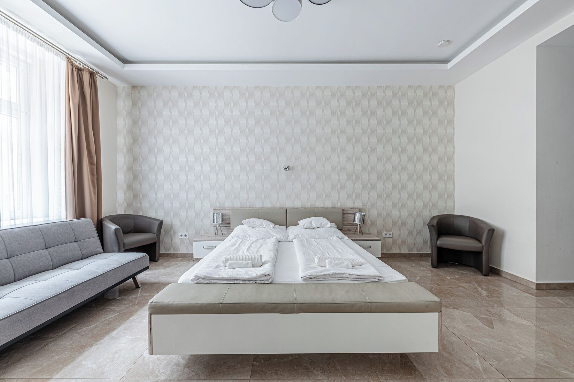Moderni hotellihuone, jossa on suuri sänky, sohva, kaksi nojatuolia ja yöpöytä, jossa on lamppu.