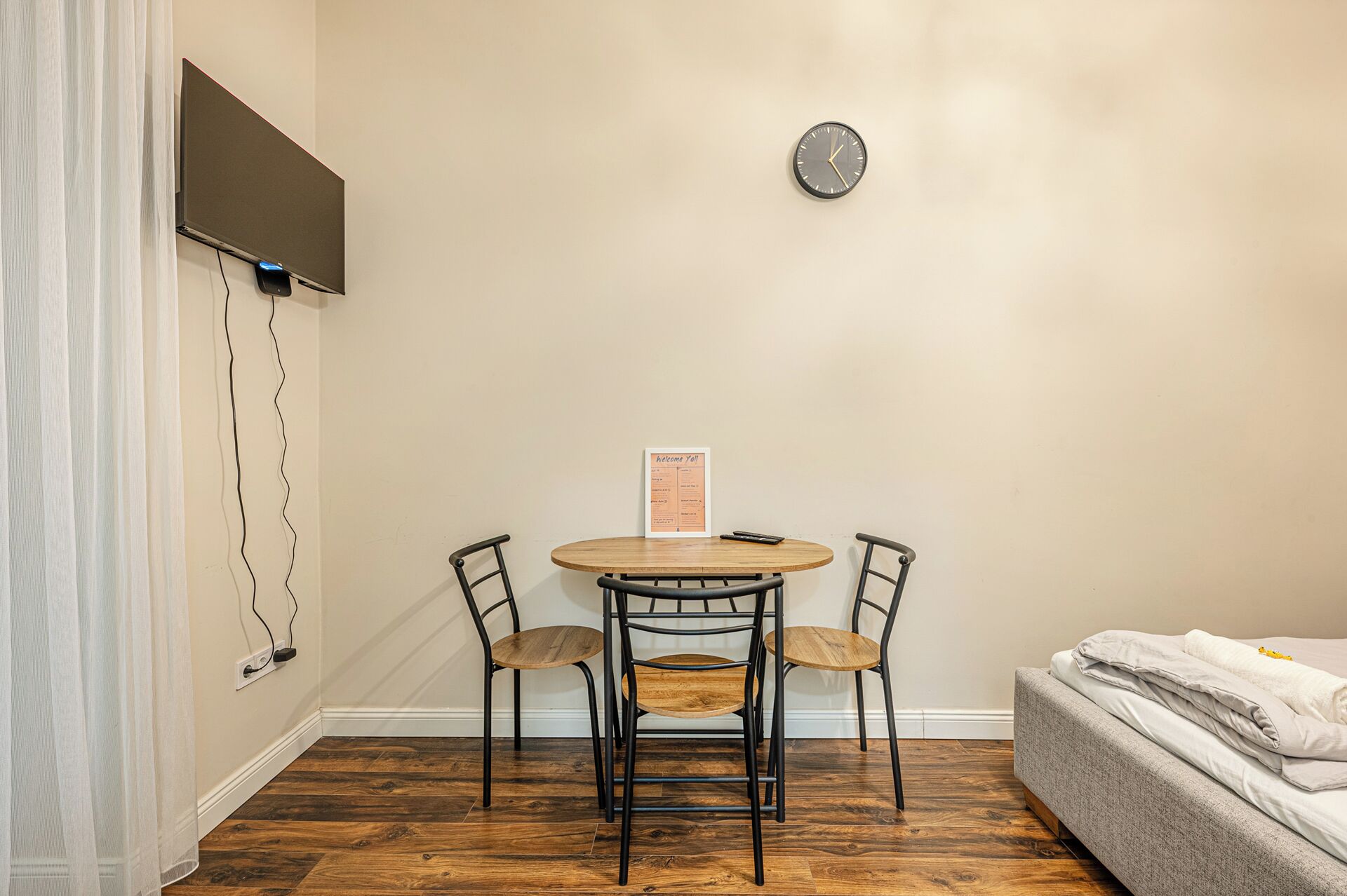Une pièce avec une table ronde en bois, deux chaises, une horloge au mur, un lit et une pancarte sur le mur avec l’inscription « Bienvenue à tous ».