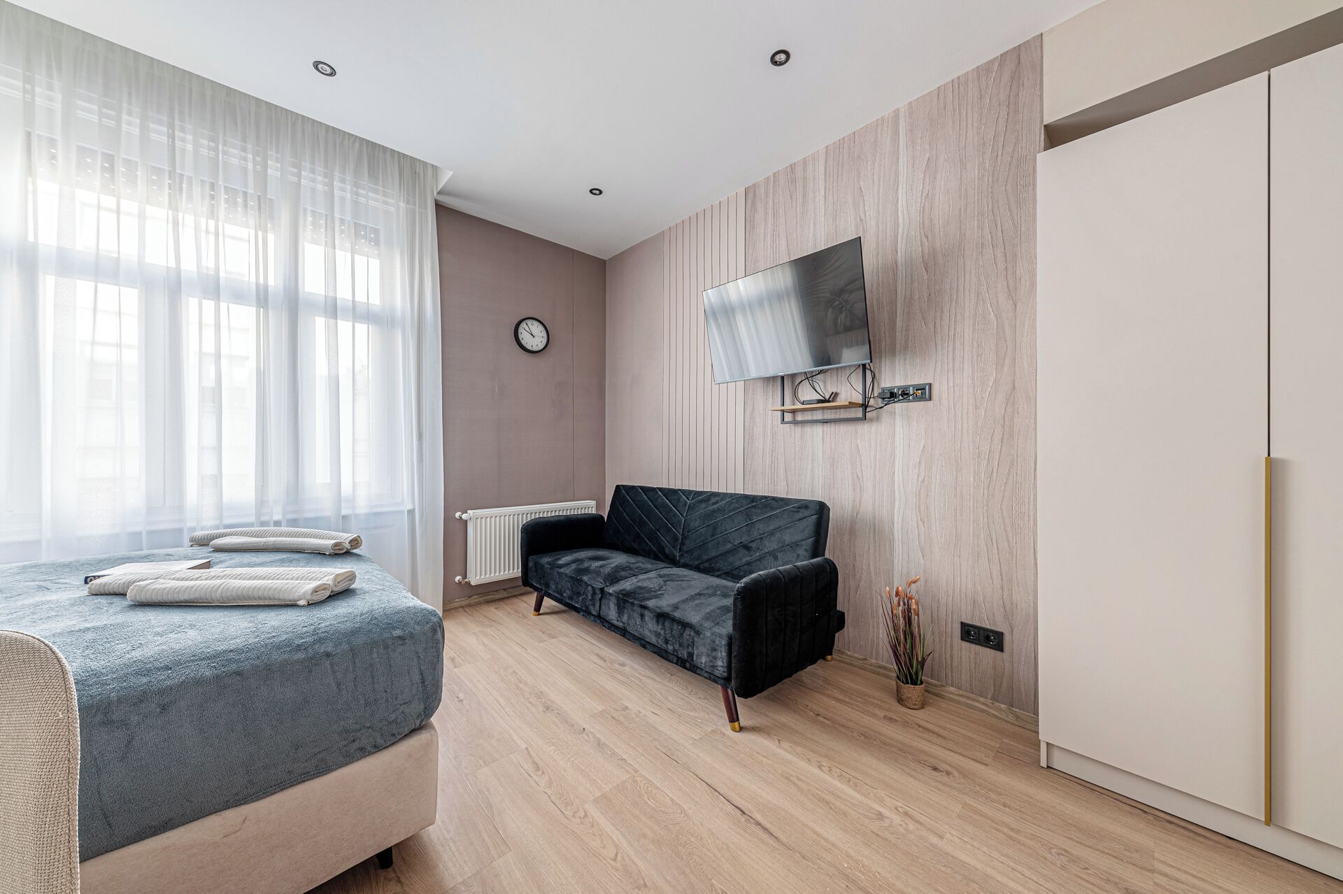 Ein modernes Schlafzimmer mit Bett, Sofa, an der Wand montiertem Fernseher und einem großen Fenster mit transparenten Vorhängen.