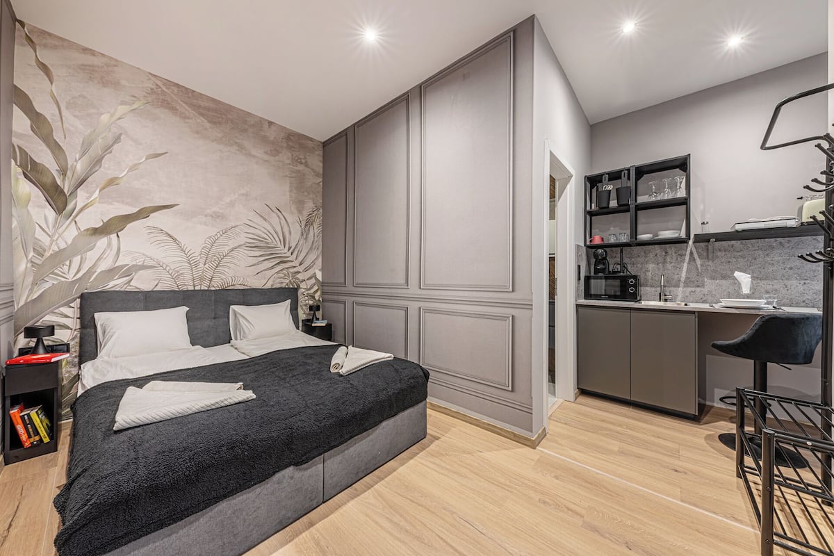 A modern bedroom with a large bed, a small desk, and a kitchenette.
