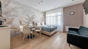 Une chambre moderne avec un lit, un bureau avec des chaises, une grande fenêtre avec des rideaux légers, un radiateur et une horloge au mur.