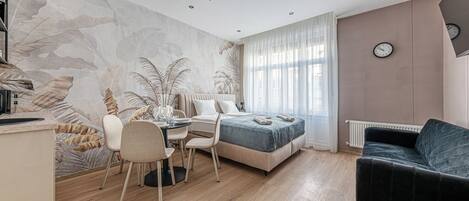 A modern bedroom with a bed, a desk with chairs, a large window with sheer curtains, a radiator, and a clock on the wall.