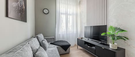 Ein modernes Wohnzimmer mit einem grauen Sofa, einem Flachbildfernseher auf einem schwarzen Standfuß, einer Uhr an der Wand und einem Fenster mit transparenten Vorhängen.