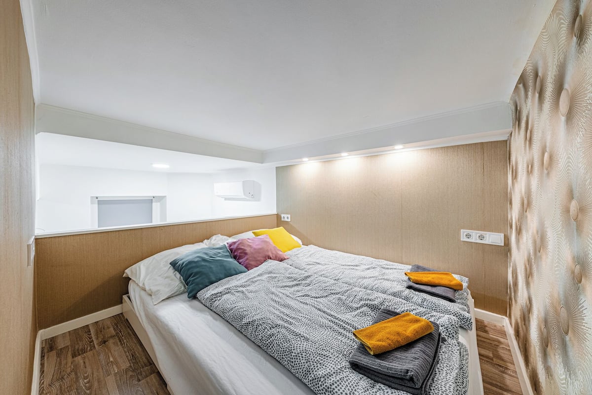 A hotel room with a bed, a mirror, a wall-mounted air conditioning unit, and a wooden floor.