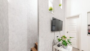 Un pasillo con un televisor montado, un banco blanco con una planta y una puerta blanca.