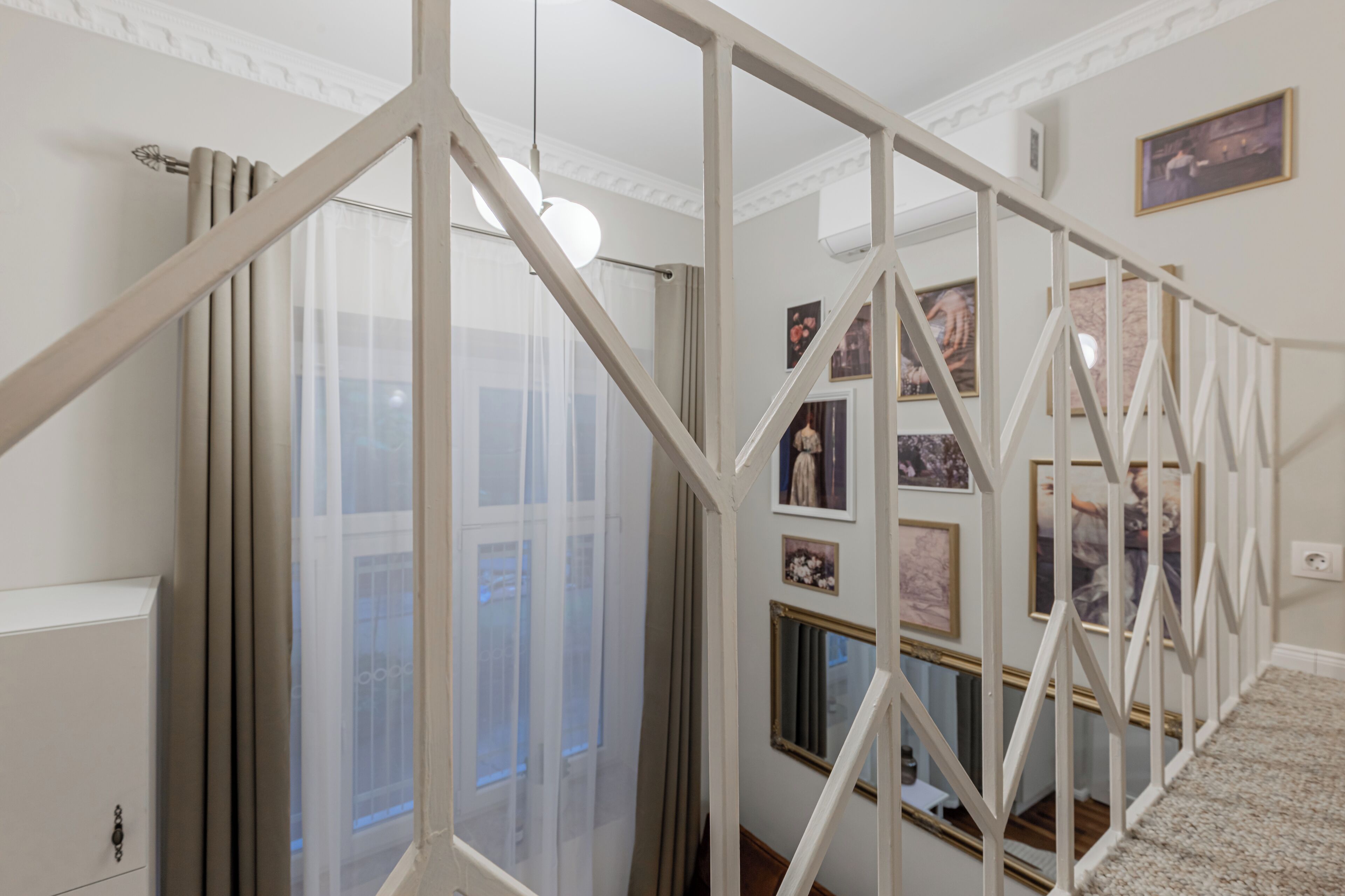Un escalier avec des rampes blanches, une fenêtre avec des rideaux légers et un mur orné de tableaux encadrés.
