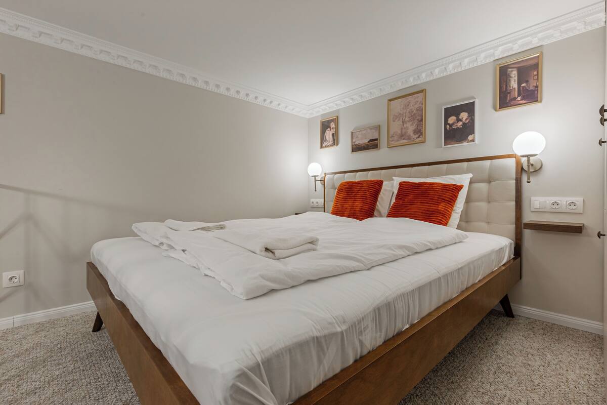 A neatly made bed with white linens, a wooden headboard, and two wall-mounted light fixtures.