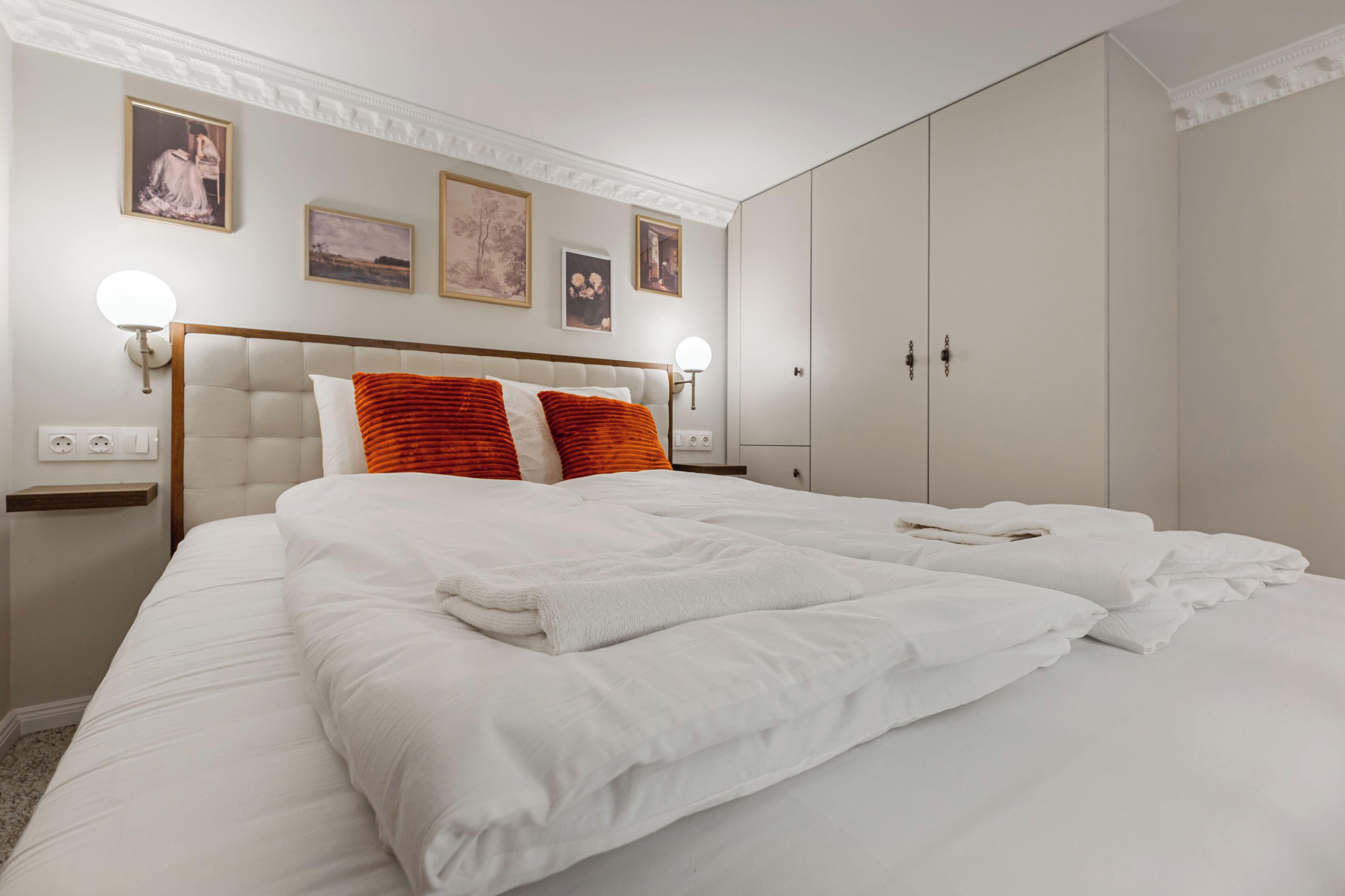 A neatly made bed with white linens, a wooden headboard, and two orange pillows. There are wall-mounted lights, a wardrobe, and framed pictures on the wall.