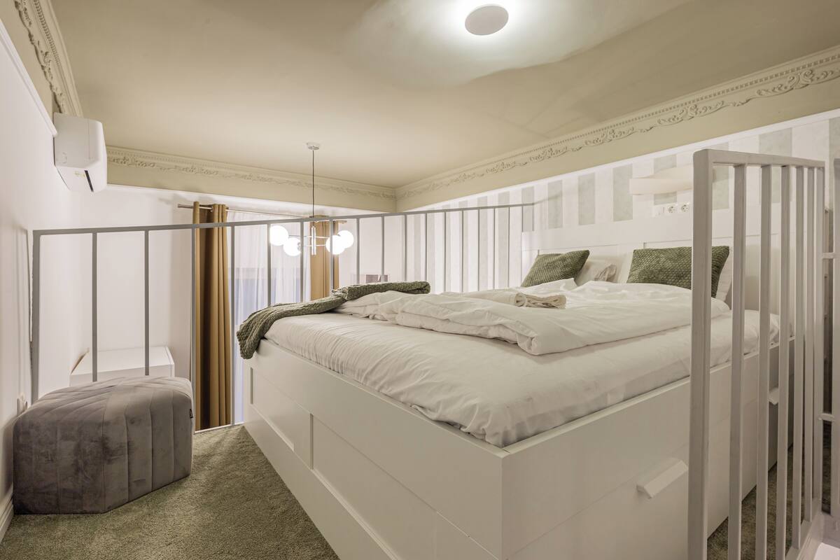 A bedroom with a white bed, a grey ottoman, and a white wardrobe.