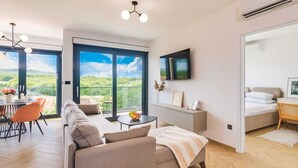 Uma sala de estar moderna com sofá, mesa de jantar e cadeiras, televisão na parede e varanda com vista panorâmica.