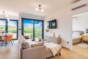 Ein modernes Wohnzimmer mit einer Couch, einem Esstisch und Stühlen, einem an der Wand montierten Fernseher sowie einem Balkon mit schöner Aussicht.