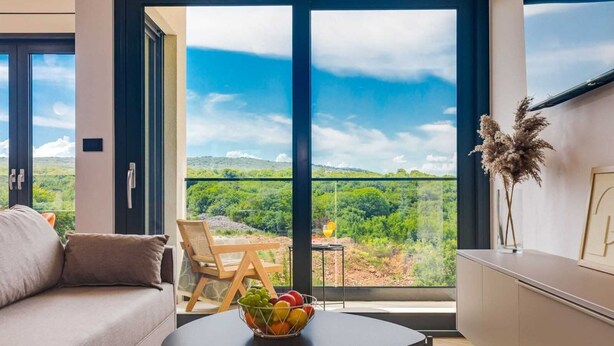 Uma sala de estar moderna com um sofá, uma mesa de jantar com frutas e vista para uma paisagem verde através de grandes janelas.