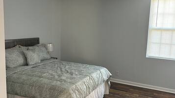A bedroom with a bed, wooden flooring, a window with blinds, and a ceiling light fixture.