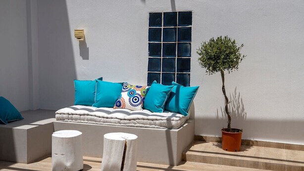 Un área de estar exterior blanca con un banco acolchado, dos taburetes blancos y una planta en maceta.