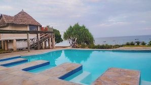 A swimming pool with stone steps, a thatched-roof building, and a view of the sea. - Coral Cove Diani Treehouses (Diani Beach)