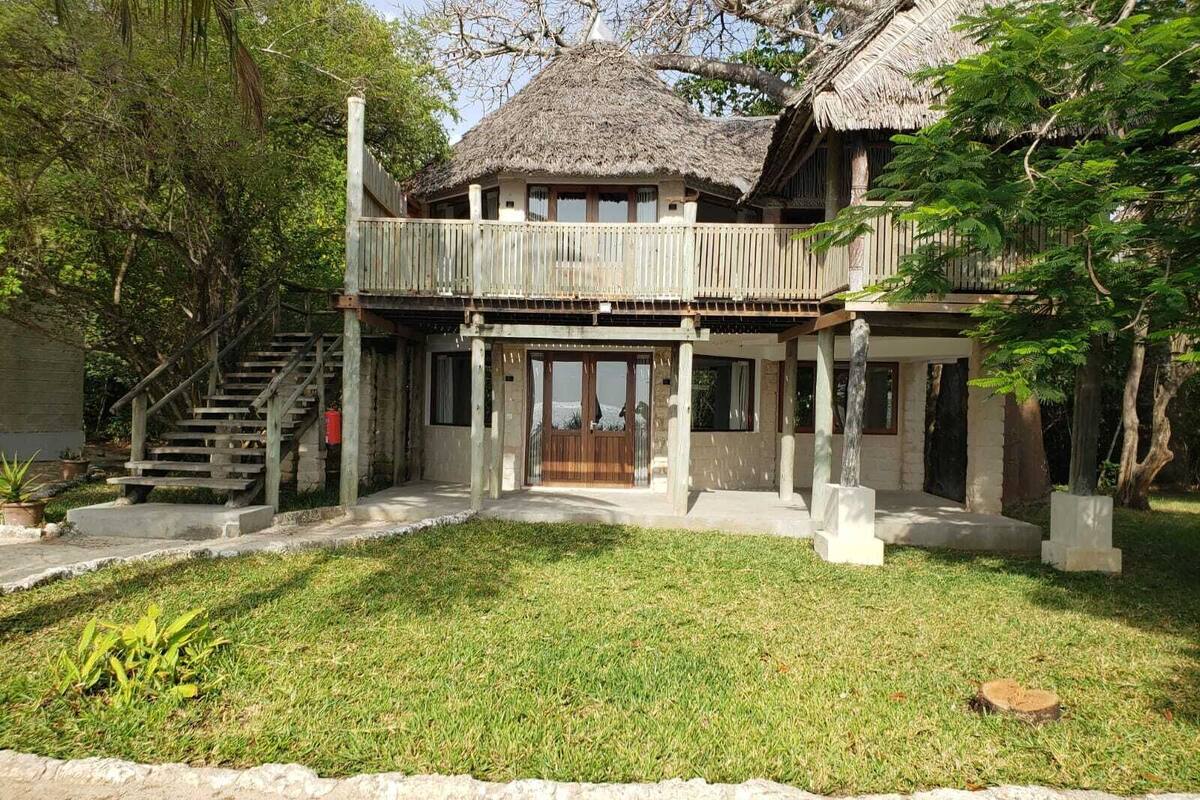 Une maison à deux étages, avec un toit de chaume, un balcon en bois et un escalier en béton.
