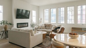 Un vaste salon comprenant un grand canapé, une table basse et une table à manger avec des chaises. On y trouve également une cheminée, un téléviseur fixé au mur et de grandes fenêtres qui laissent entrer la lumière naturelle.