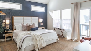 Une chambre à coucher avec un grand lit, une tête de lit en bois, une table de chevet et une commode. Il y a deux fenêtres avec des rideaux, une vue sur l’extérieur et un balcon avec une balustrade.