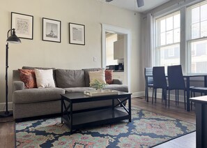 A living room with a grey sofa, a black coffee table, and a patterned rug. There are framed pictures on the wall, a dining area with chairs, and a kitchen in the background. - The Harrison C - KING 1BD/1BA Apt near Riverfront (Evansville)