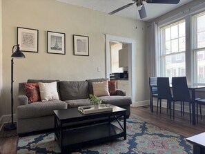 A living room with a grey sofa, a black coffee table, and a patterned rug. There are framed pictures on the wall, a ceiling fan, and a view of the kitchen through an open door. - The Harrison C - KING 1BD/1BA Apt near Riverfront (Evansville)