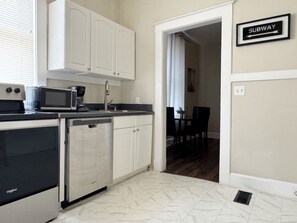 A compact kitchen with white cabinets, a dishwasher, a microwave, and a coffee maker. A sign indicating a subway direction is visible on the wall. - The Harrison C - KING 1BD/1BA Apt near Riverfront (Evansville)