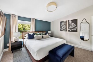 Un dormitorio ordenado con cama, mesitas de noche, un banco, una ventana con cortinas y una obra de arte colgada en la pared.