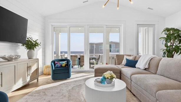 A spacious living room with a large sofa, a coffee table, and a television mounted on the wall. There is a sliding glass door leading to an outdoor patio area with a view of the ocean. - Fall Availability | Steps to Beach | 7 bdrm home (Holden Beach)