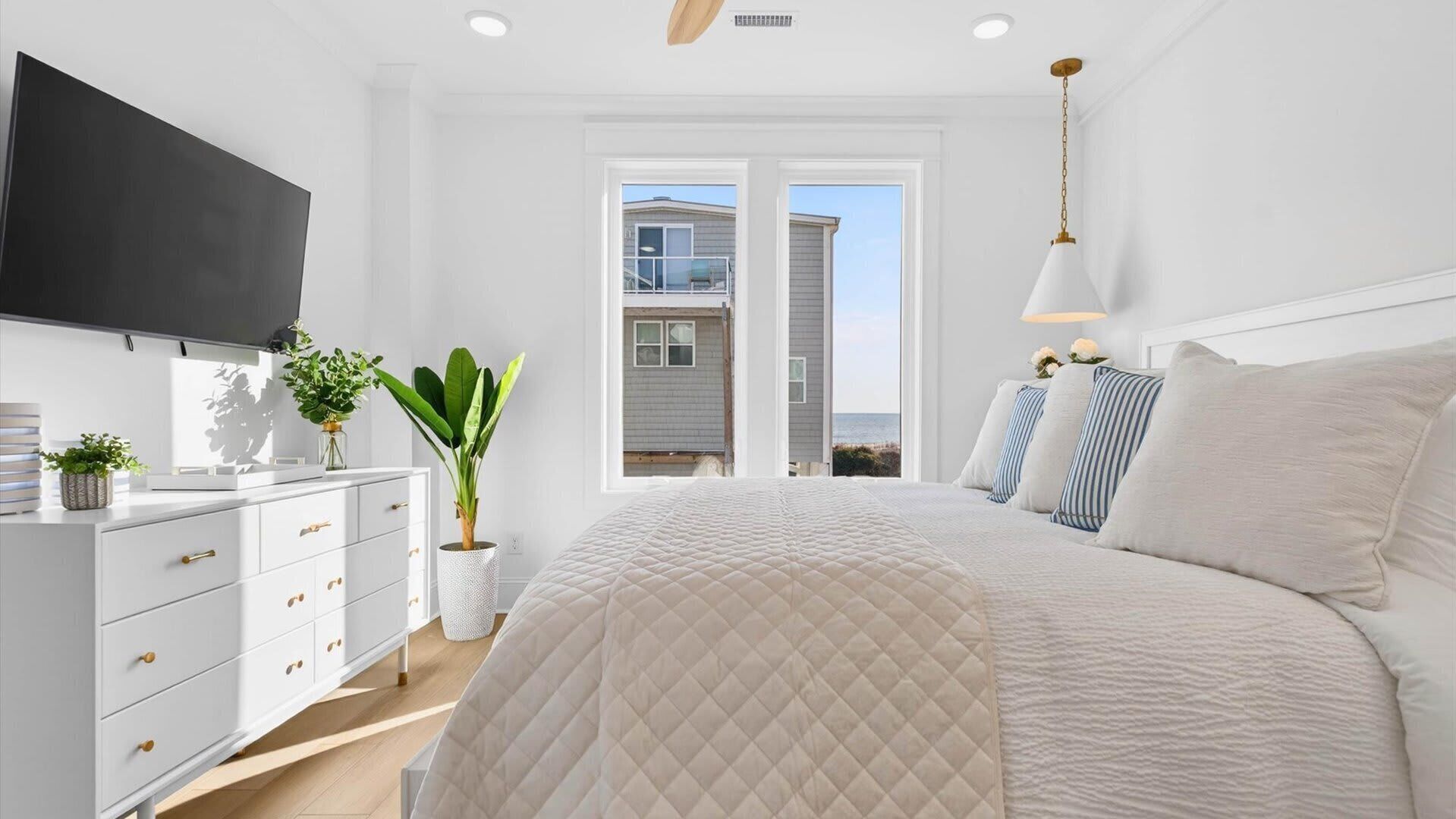 A bedroom with a large bed, a TV mounted on the wall, a dresser, and a potted plant.