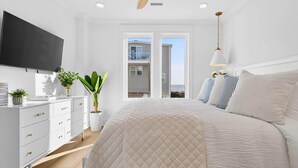A bedroom with a large bed, a TV mounted on the wall, a dresser, and a potted plant. - Fall Availability | Steps to Beach | 7 bdrm home (Holden Beach)
