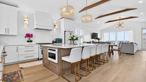 A modern kitchen with white cabinetry, a central island with bar stools, pendant lighting, and a view to the outside. - Fall Availability | Steps to Beach | 7 bdrm home (Holden Beach)