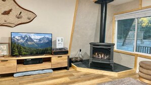 A cozy living room with a wooden entertainment unit, a flat-screen TV, a fireplace, and a window with a view of a deck and trees. - Adventure's Retreat for Trails & Forest Fun! (Overgaard)
