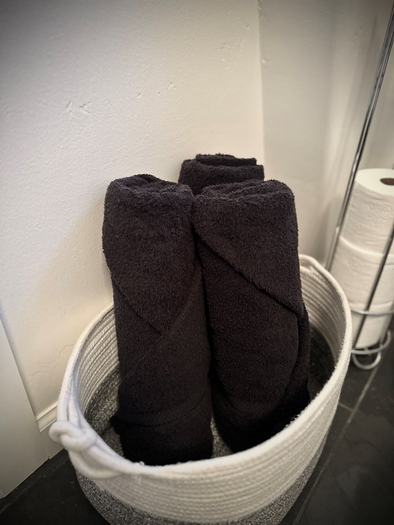 A white laundry basket with dark towels inside, placed against a white wall.