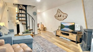 A modern living room with a staircase, a flat-screen TV, a fireplace, and a decorative wall piece. - Adventure's Retreat for Trails & Forest Fun! (Overgaard)