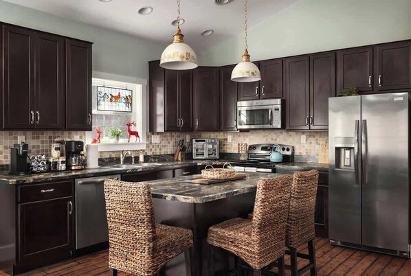 A modern kitchen with dark cabinets, stainless steel appliances, a central island with bar stools, and pendant lighting. - Creekside Mariposa Cottage · Hot Tub & Fire Pit (Berkeley Springs)