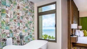 Een badkamer met een vrijstaand bad, een groot raam met uitzicht op zee, en een bed met nachtkastje.