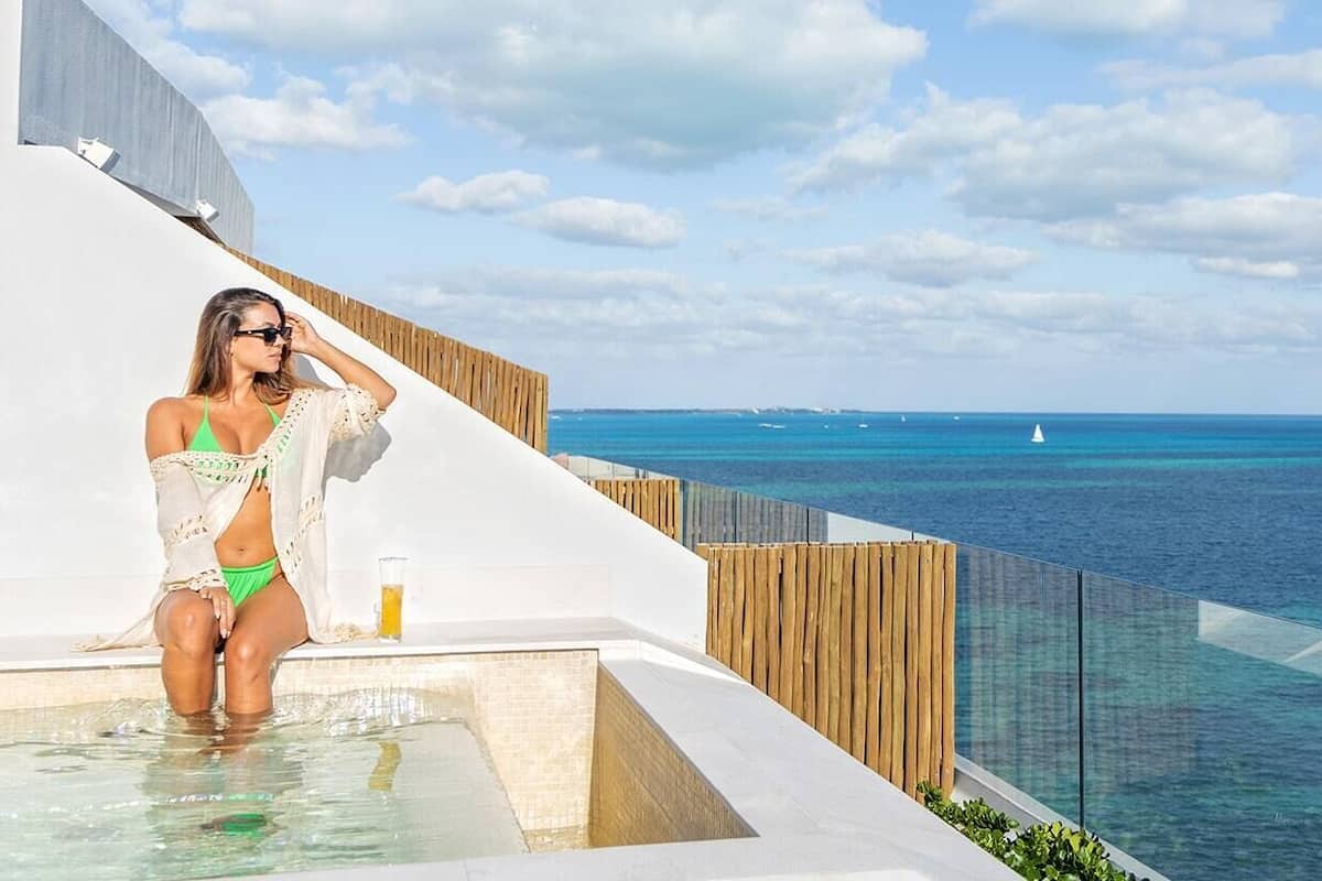 Une femme vêtue d’un bikini vert est assise au bord d’une piscine, avec vue sur l’océan et les bateaux.