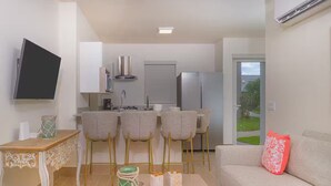 Un espace de vie compact comprenant un coin cuisine, un espace repas avec des tabourets de bar, un canapé agrémenté d’un coussin décoratif et un téléviseur à écran plat fixé au mur.
