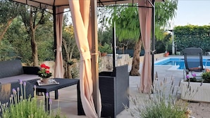 Un patio al aire libre con zona de estar cubierta, piscina y vegetación circundante.