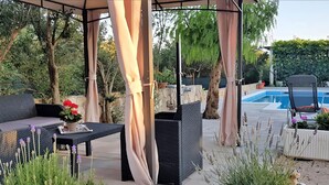 Un espace extérieur aménagé en patio, avec un coin salon couvert, une piscine et diverses plantes.